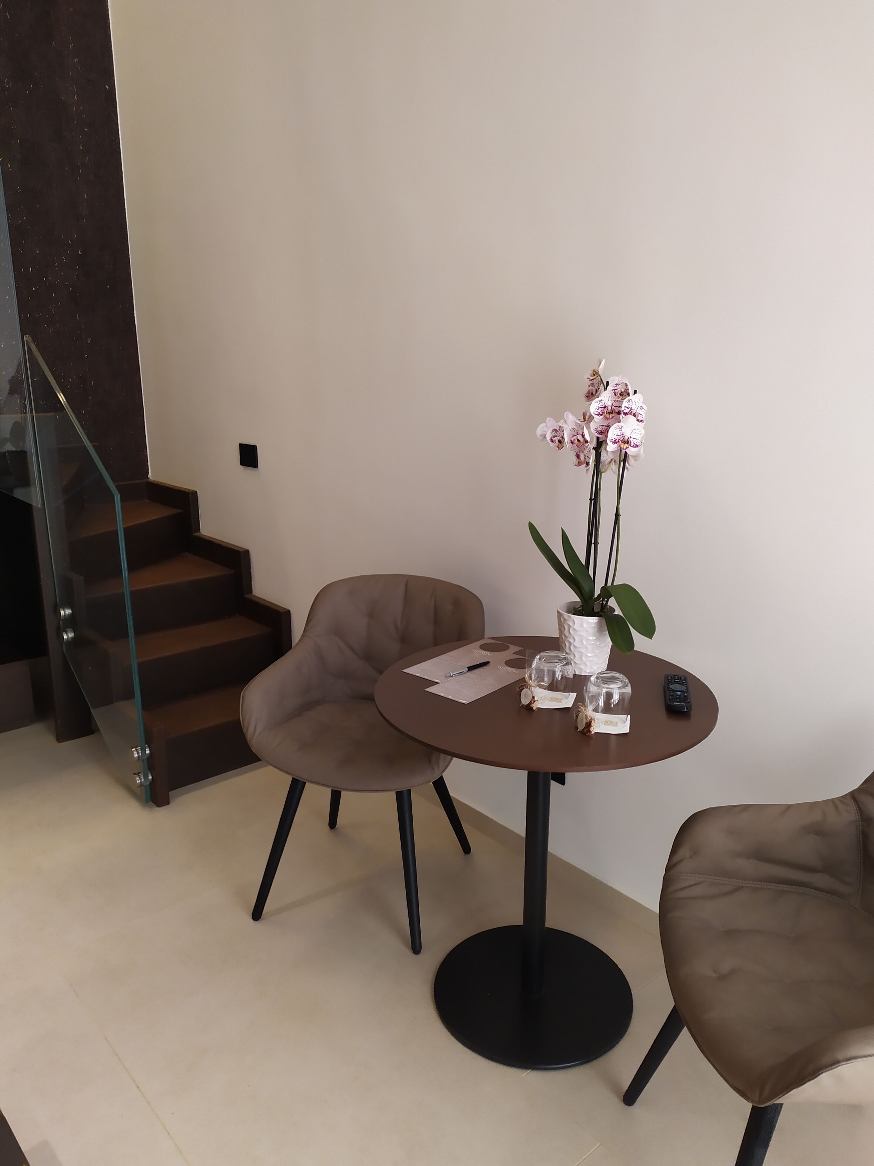 A modern living room with a round table, two chairs, a vase with orchids, and a staircase.