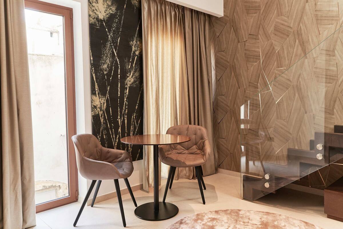 A modern dining area with a round table and two chairs, a large window with curtains, and a geometric patterned wall.