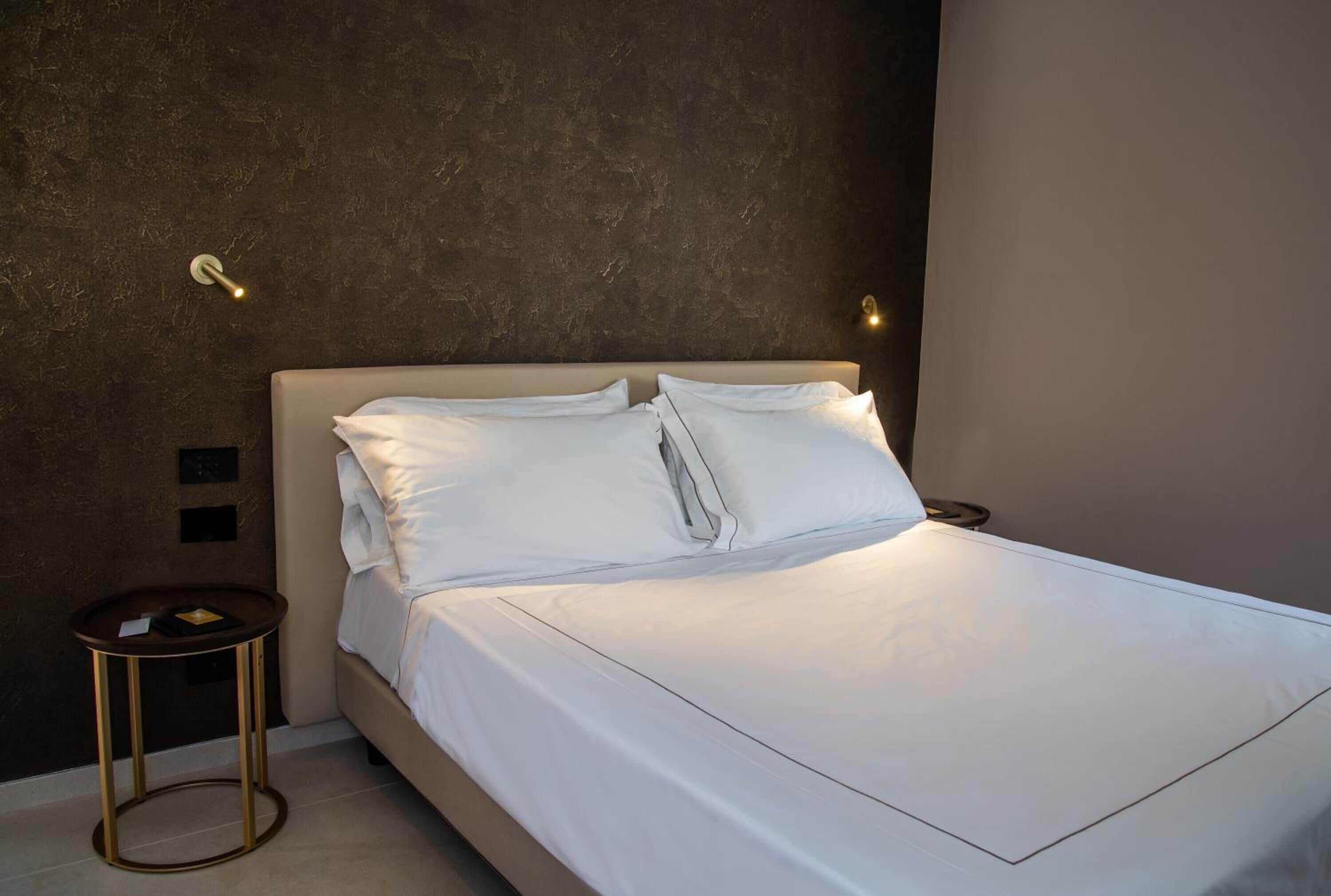 Una cama bien hecha con sábanas blancas, una mesita de noche redonda con marco dorado y una pared texturizada detrás de la cama.