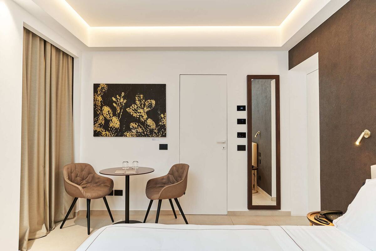 A modern hotel room with a bed, two chairs, a small table, a painting, a door, and a mirror.