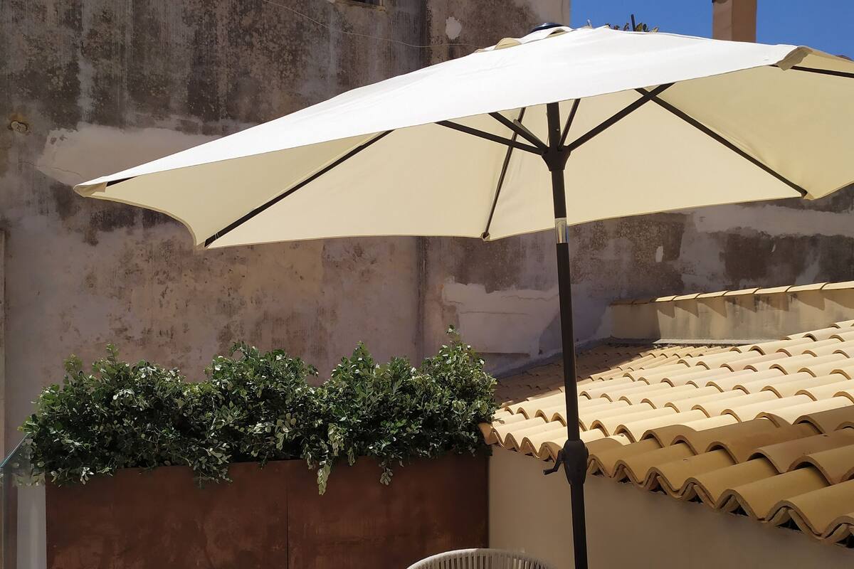 A patio with a table and chairs under a large umbrella, surrounded by a low wall with greenery and a tiled roof above.