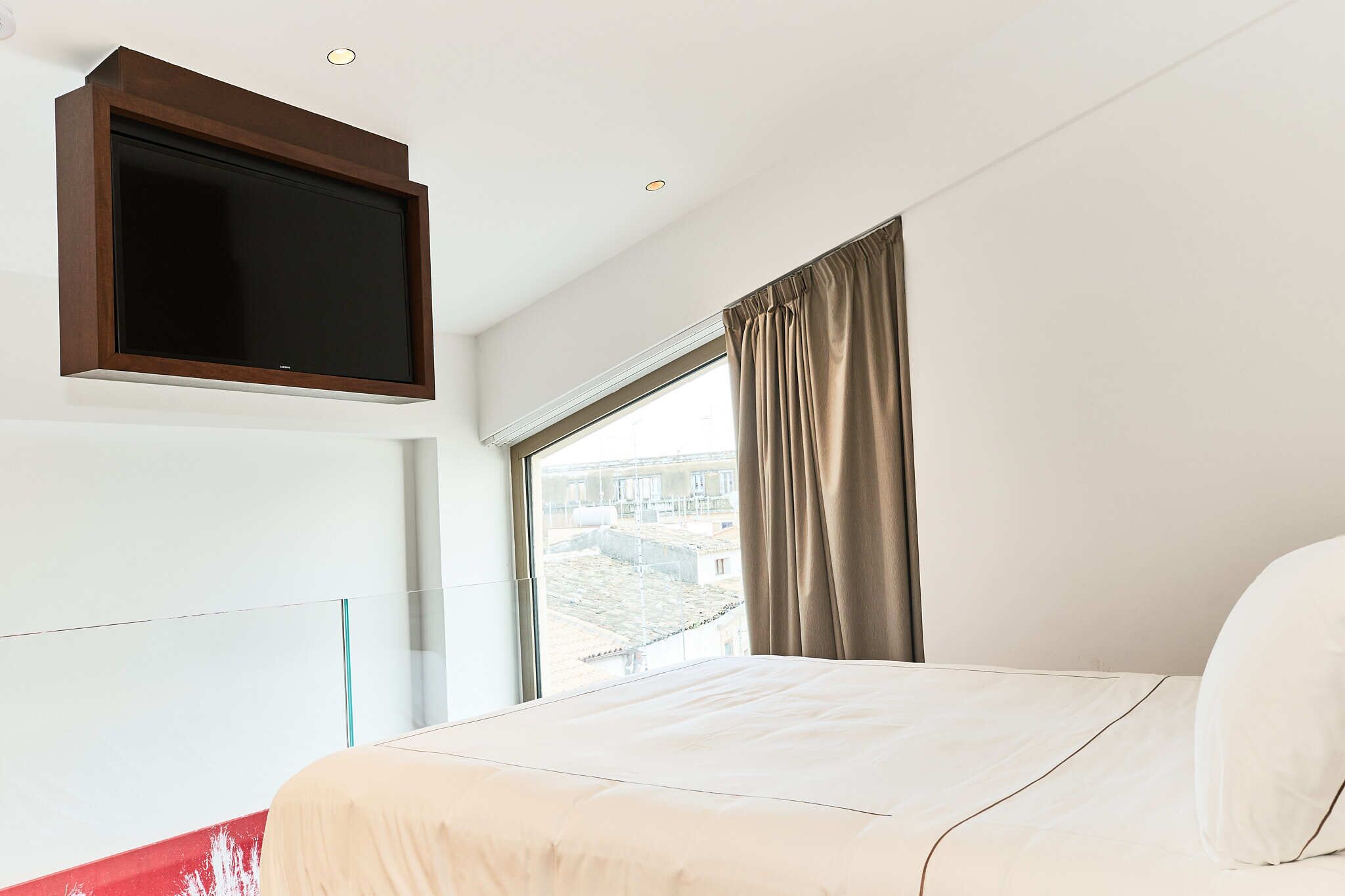 A hotel room with a large bed, a mounted TV, a window with curtains, and a view of buildings outside.