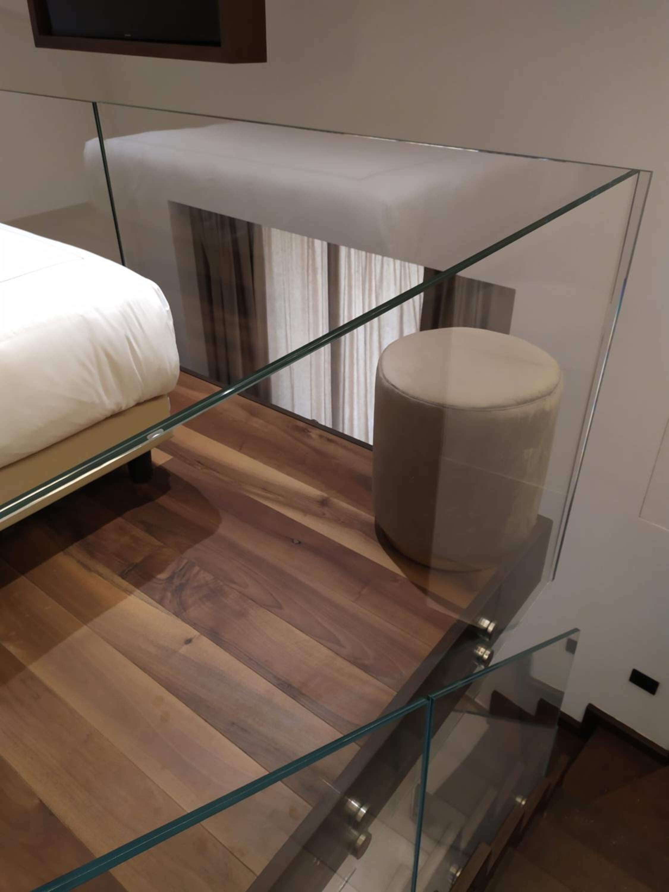 Un dormitorio moderno con una escalera de cristal, suelo de madera, una cama y un reposapiés redondo.