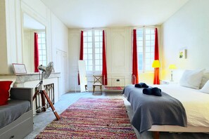 A hotel room with a bed, a desk, a chair, a standing fan, a red curtain, a yellow lamp, and a colorful rug.