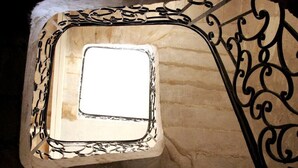 Una ventana cuadrada con un marco de metal decorativo, vista a un muro de piedra y una barandilla de metal con diseños intrincados.
