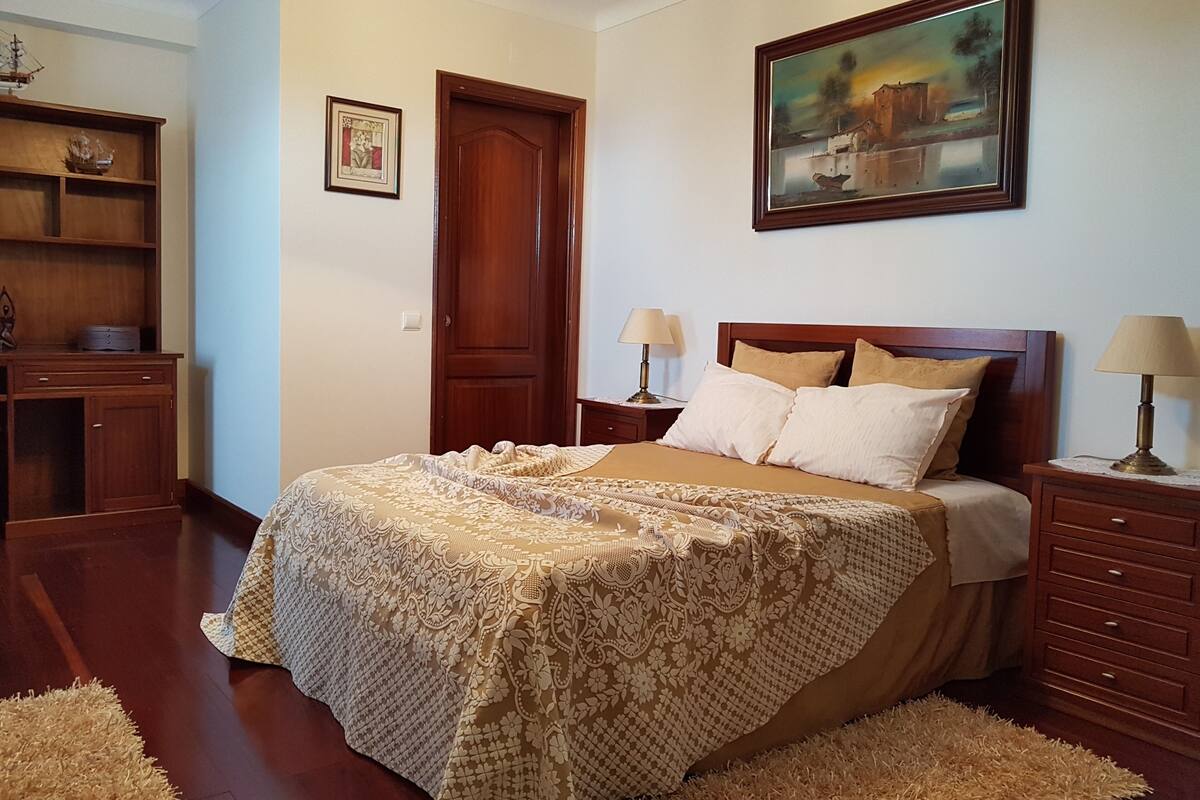 A bedroom with a large bed, wooden headboard, two bedside tables with lamps, a wooden wardrobe, a framed painting on the wall, and a door.