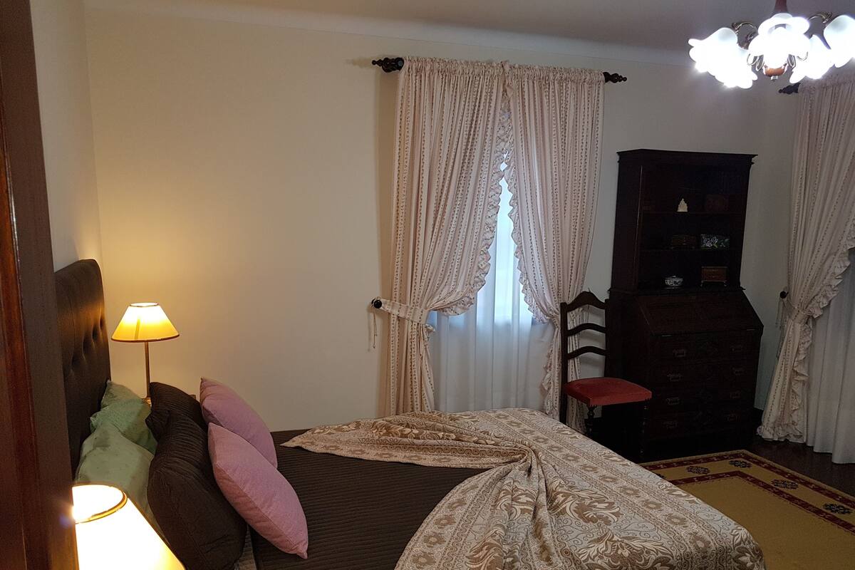 A bedroom with a bed, a nightstand, a lamp, a chair, a dresser, and a window with curtains.