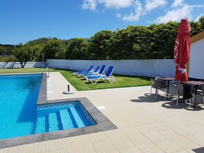 A swimming pool with blue water, surrounded by a tiled deck. There are blue lounge chairs and a red umbrella. - Casa Marques (Angra do Heroismo)