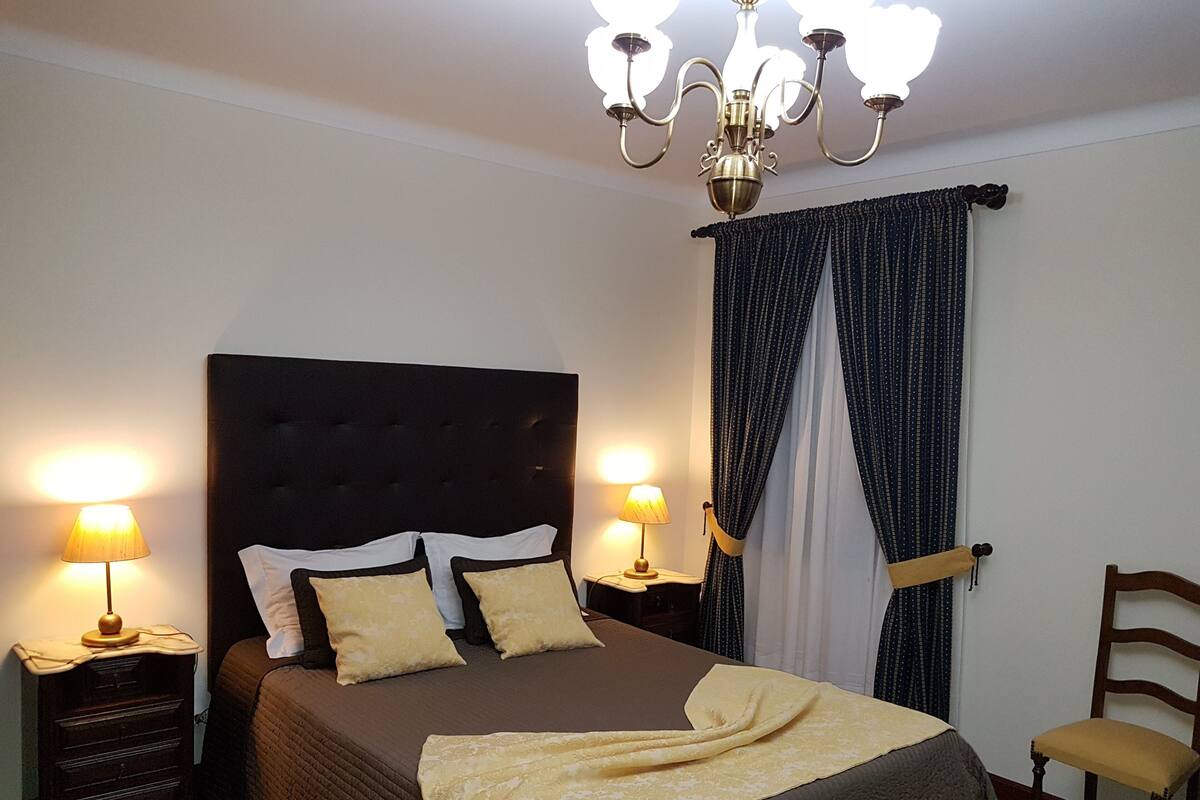A neatly made bed with a dark headboard, two bedside lamps, a wooden chair, and a chandelier.