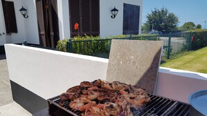 A barbecue grill with grilled chicken in front of a white building with shutters and a green fence. - Casa Marques (Angra do Heroismo)