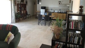 Un salon avec un canapé, une table à manger et des chaises. On y trouve une bibliothèque remplie de livres et une plante en pot. On aperçoit une cuisine à l’arrière-plan, avec un four à micro-ondes et des placards.