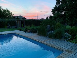 Una piscina rectangular amb un solàrium de fusta, envoltada de vegetació exuberant i un cobert petit.