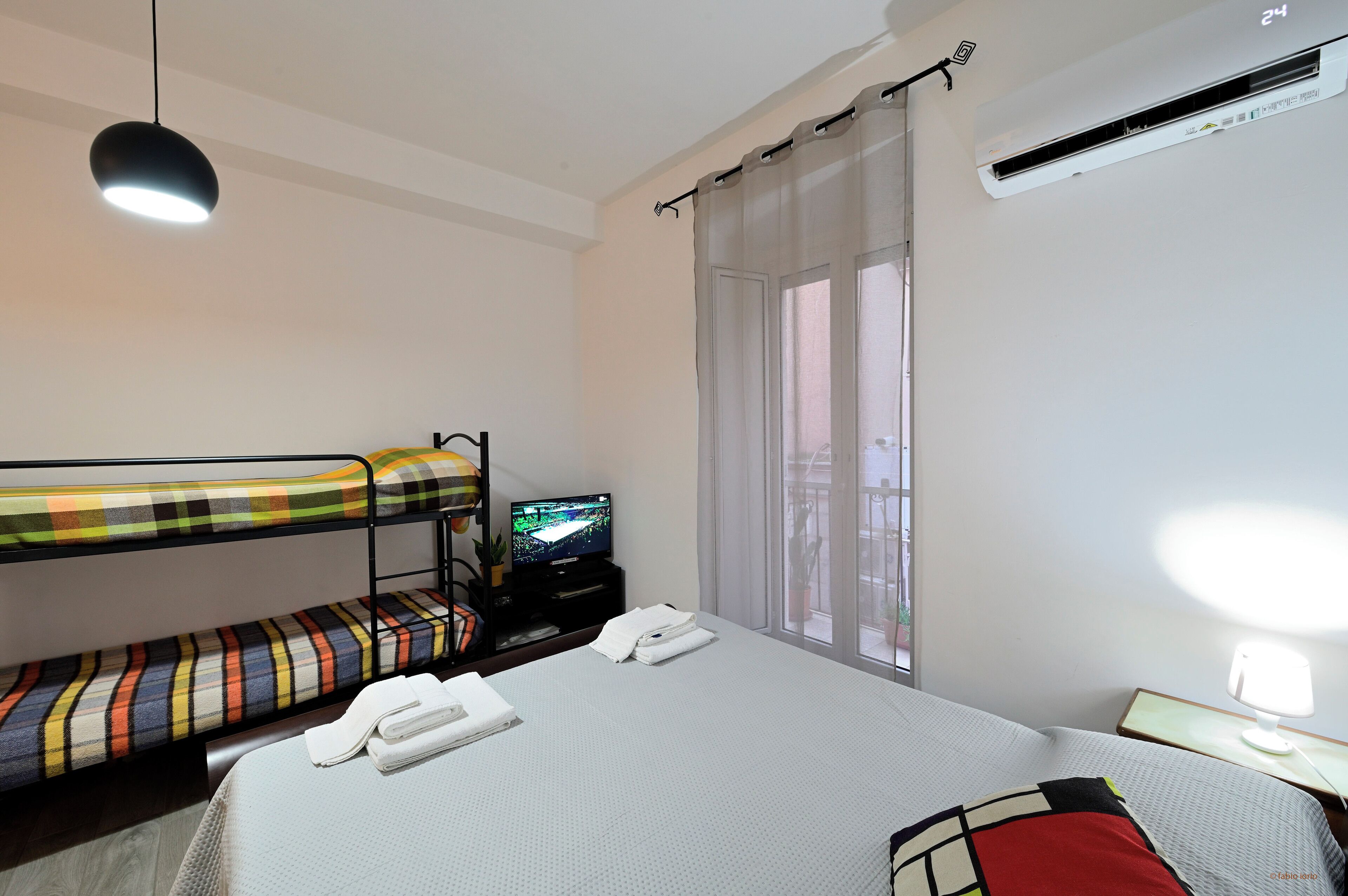 Ein Hotelzimmer mit Etagenbett, einem Einzelbett, einem Fernseher, einem Schreibtisch mit Lampe und einem fenster mit Vorhängen.