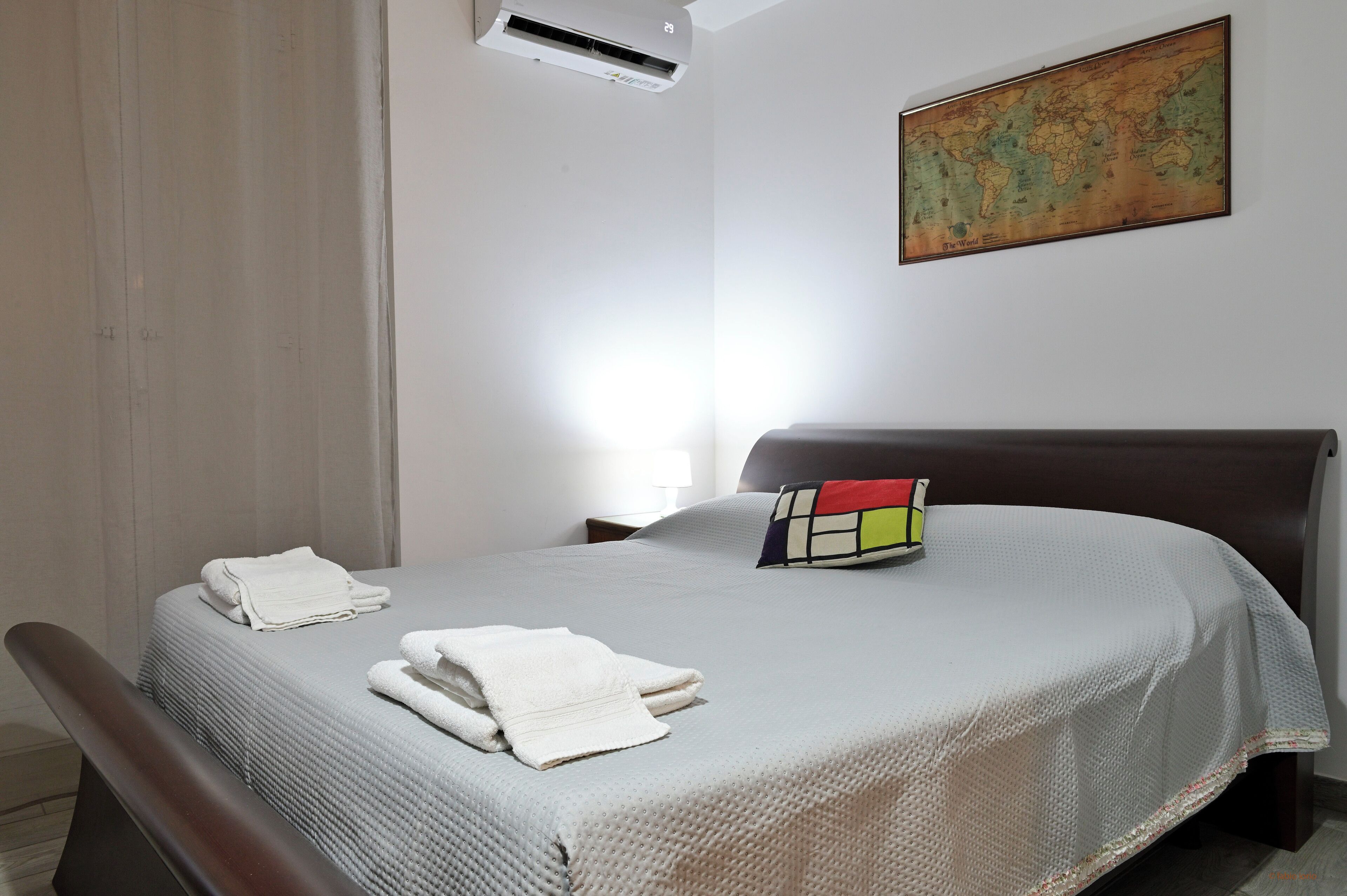 A neatly made bed with a white blanket and two folded towels, a colorful pillow, a world map on the wall, and an air conditioning unit above the bed.