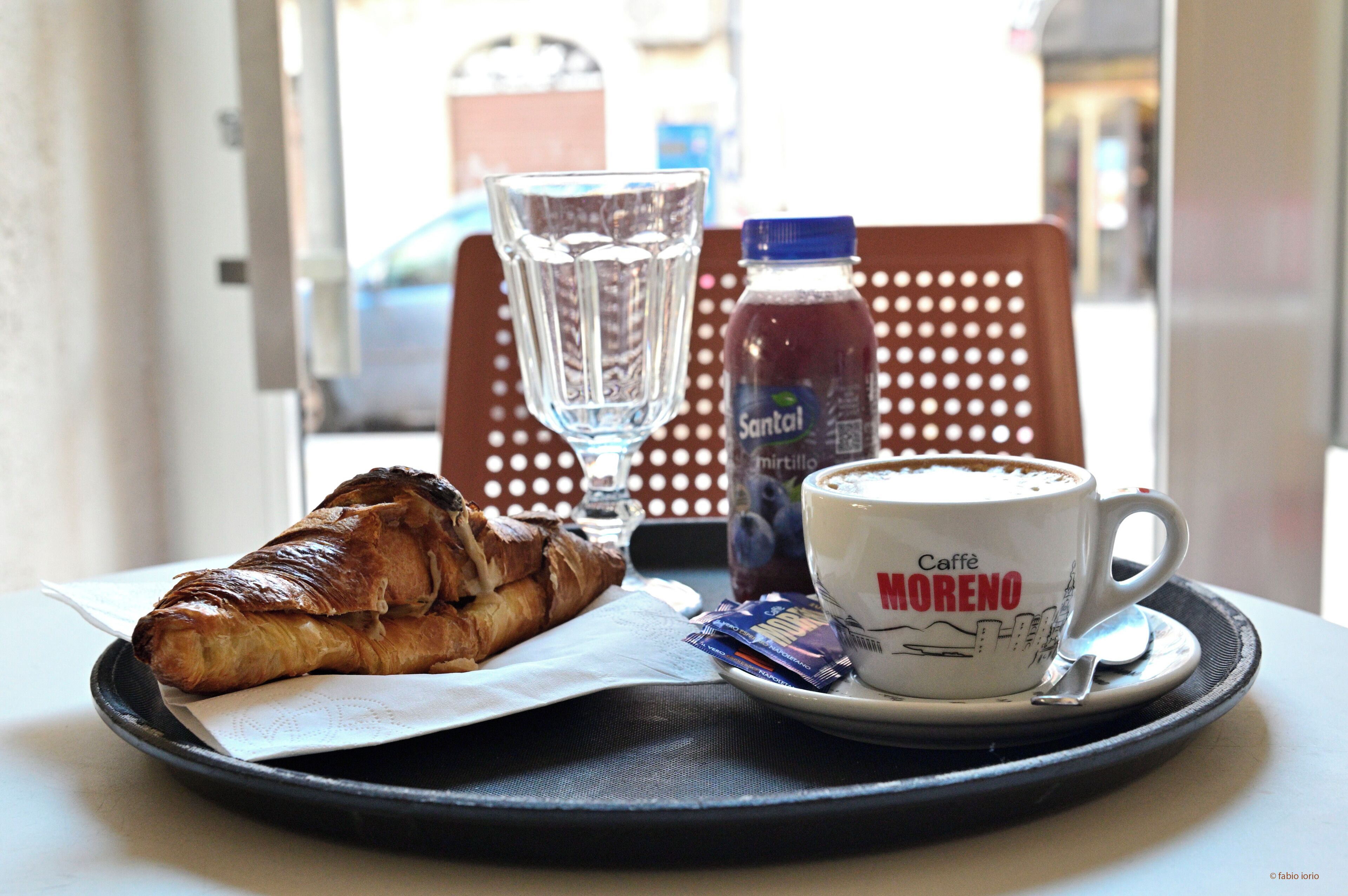 A tray with a croissant, a glass of water, a bottle of Santal, and a cup of coffee labeled "Caffè MORENO".