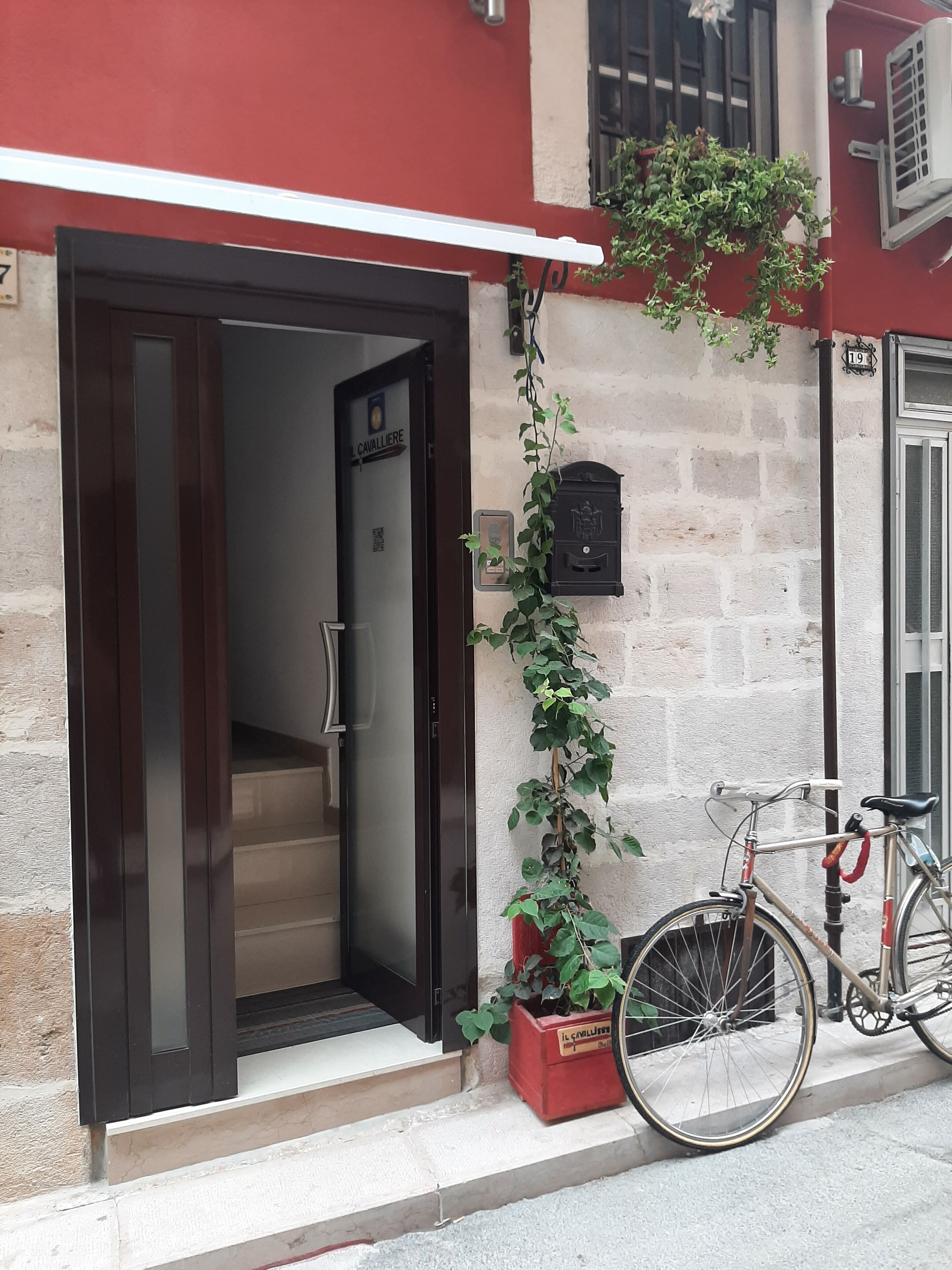 Entrada de un edificio con puerta de cristal, una escalera que conduce al interior, un buzón rojo y una bicicleta estacionada afuera.