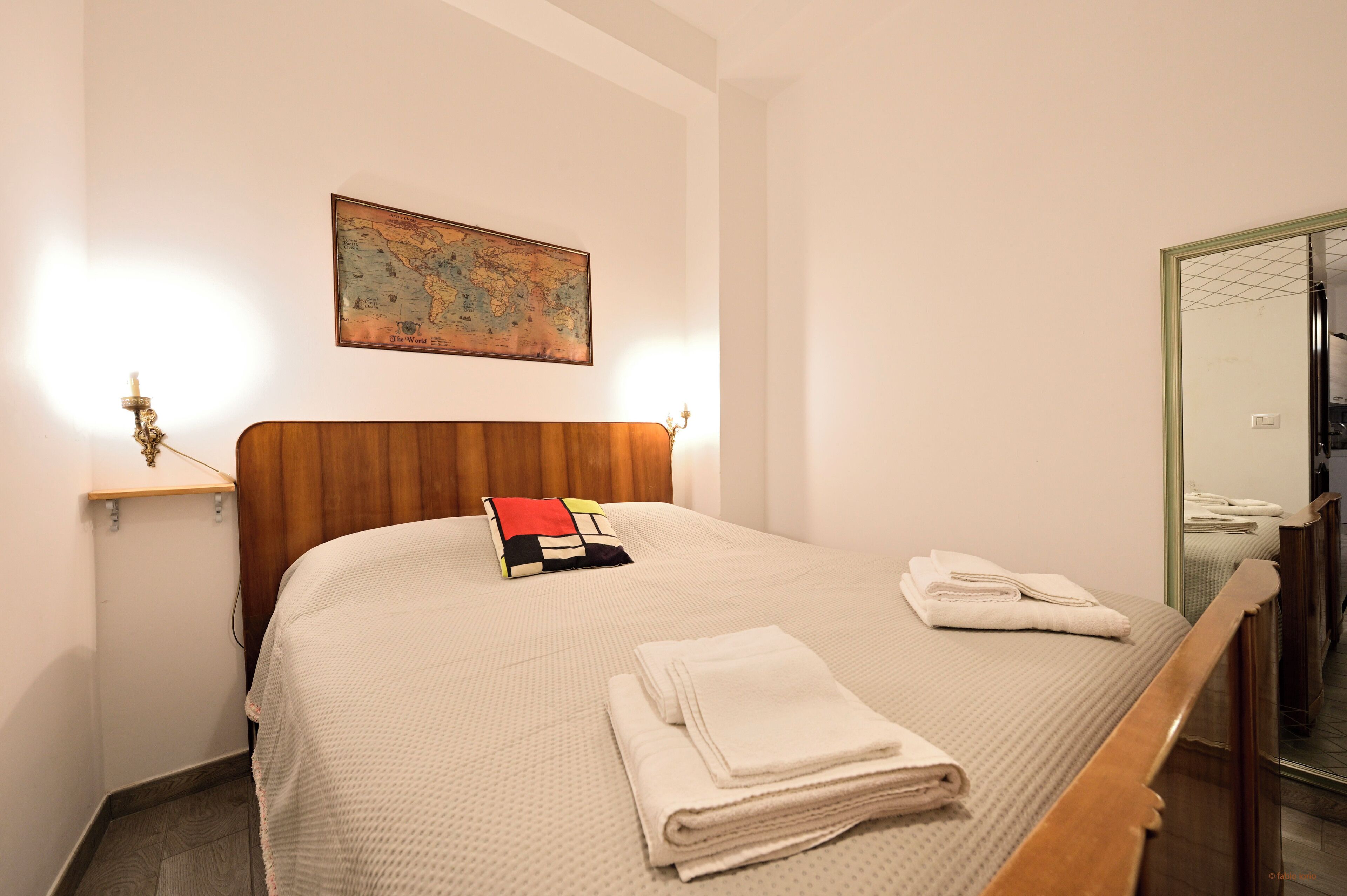 A neatly made bed with a wooden headboard, a map on the wall, and a mirror reflecting part of the room.