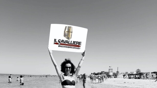 A woman standing on a beach holding a sign that reads "IL CAVALLIERE BEDANDBREAKFAST". - Il Cavalliere Bed & breakfast (Barletta)