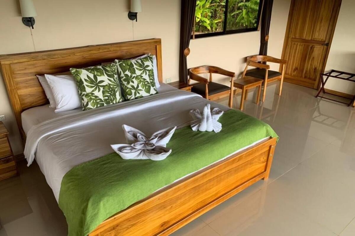A neatly made bed with a green bedspread and two towels, wooden headboard, and a wooden chair in a well-lit room with a large window.