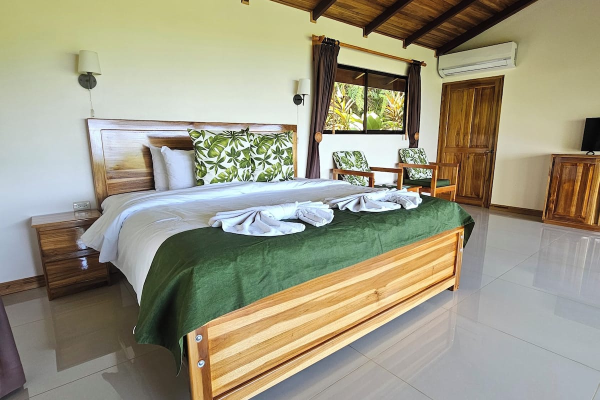 A spacious bedroom with a wooden bed, a green throw, a ceiling fan, and a window with curtains.