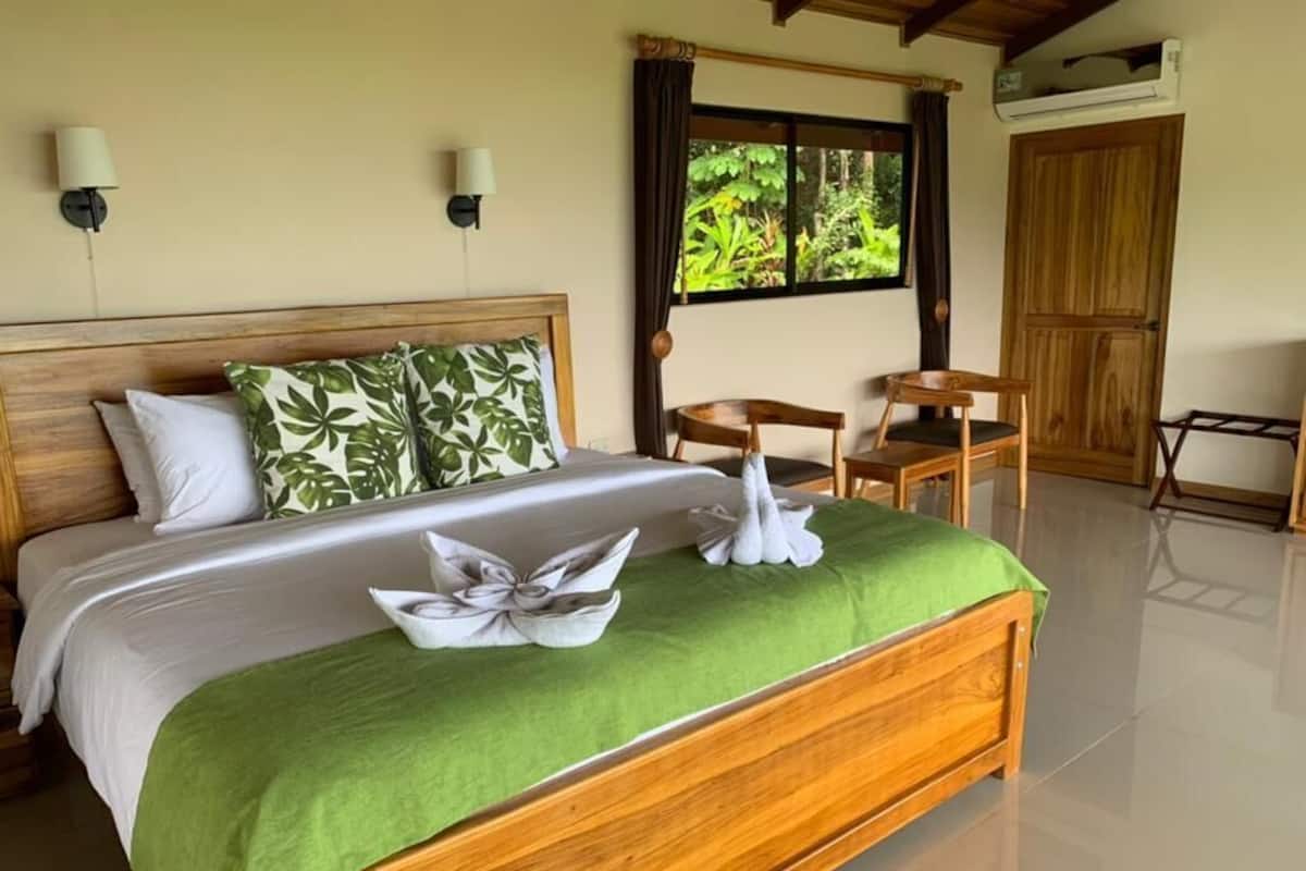 A neatly made bed with a green bedspread and white pillows, a wooden headboard, a wooden nightstand, a small wooden table, and a chair.