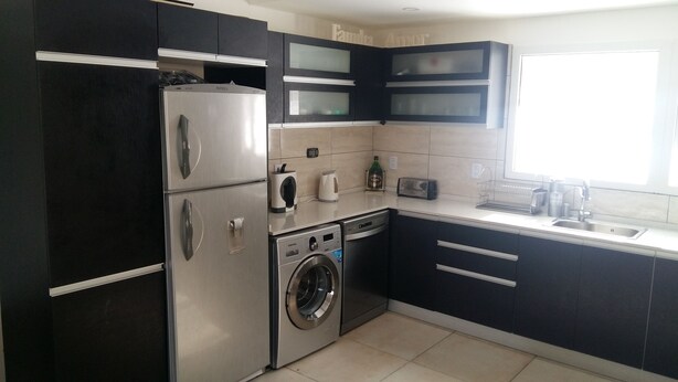 A compact kitchen with a stainless steel refrigerator, a built-in washing machine, and a sink with a window above it.