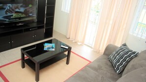 A living room with a flat-screen TV on a black entertainment unit, a glass coffee table with magazines, a grey sofa with a striped pillow, and a red and beige rug. - Crown Point Condo (Crown Point)