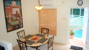 A dining area with a round table, chairs, a hanging light fixture, a painting, a window with blinds, and a view of a pool through a door. - Crown Point Condo (Crown Point)