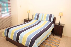 A bed with striped bedding, two bedside tables with lamps, a window with blinds, and a patterned rug.