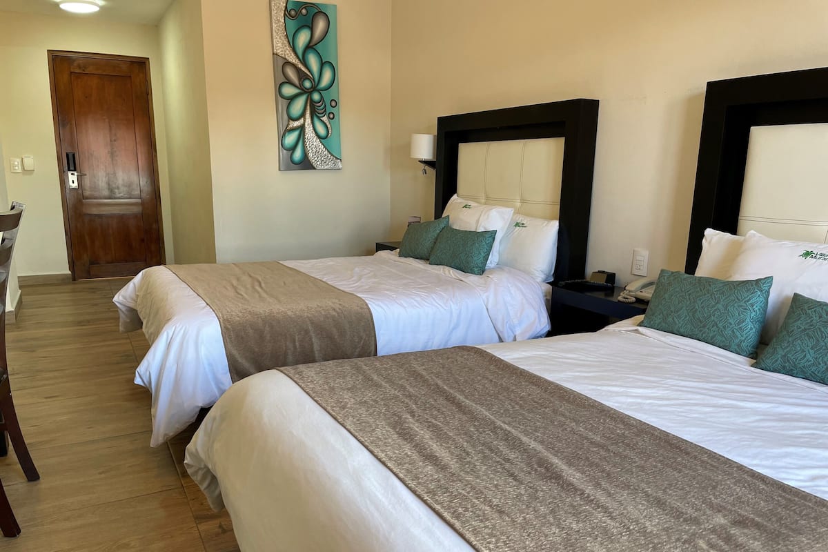 A hotel room with two beds, each with white linens and green pillows, a wooden door, a wall art piece, and a nightstand with a lamp.