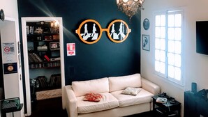 Een kamer met een donkerblauwe accentmuur, een witte leren bank, een kroonluchter, een boekenkast en een raam met jaloezieën.