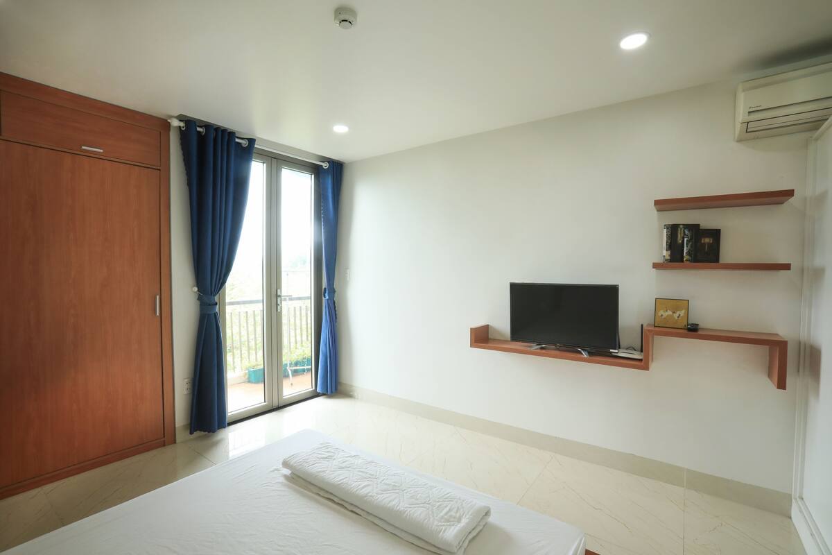 Un dormitorio con cama, armario, televisor en estante y balcón con cortinas azules.