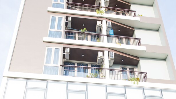 Un edificio de varios pisos con entrada de vidrio, mesas al aire libre y balcones con plantas.