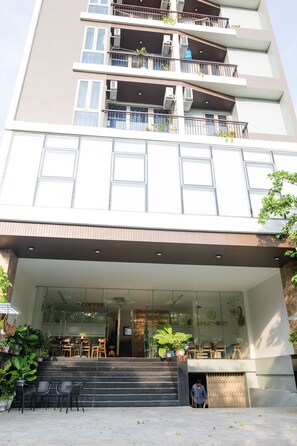 Un edificio de varios pisos con entrada de vidrio, mesas al aire libre y balcones con plantas.