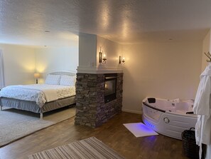 A bedroom with a stone fireplace, a bed with a white comforter, a nightstand with a lamp, and a white bathtub with a towel. - Stone Hollow (Vernal)