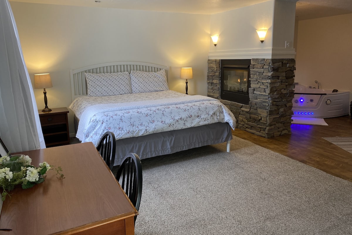 A bedroom with a bed, bedside lamps, a stone fireplace, and a spa tub.