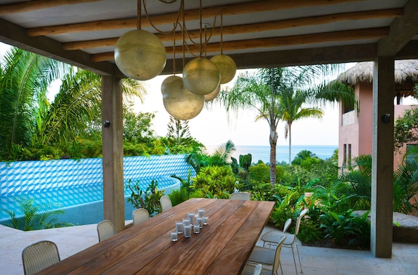 A wooden table with chairs and hanging spherical light fixtures in a covered outdoor area with a pool and lush greenery. - Designer home, Heated pool, great Ocean views close to beach and town center (Sayulita)
