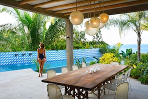 A wooden dining table with chairs, a pool with a blue and white patterned wall, and palm trees in the background. - Designer home, Heated pool, great Ocean views close to beach and town center (Sayulita)