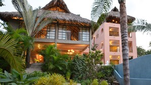 A multi-story building with a thatched roof, surrounded by tropical vegetation and a staircase leading to the entrance. - Designer home, Heated pool, great Ocean views close to beach and town center (Sayulita)