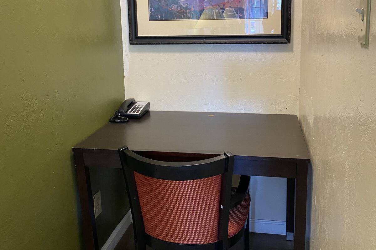 A framed landscape painting above a dark wooden desk with a red chair, a phone, and a light switch on the wall.