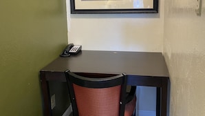 A framed landscape painting above a dark wooden desk with a red chair, a phone, and a light switch on the wall. - Holiday Motel (Morgan Hill)