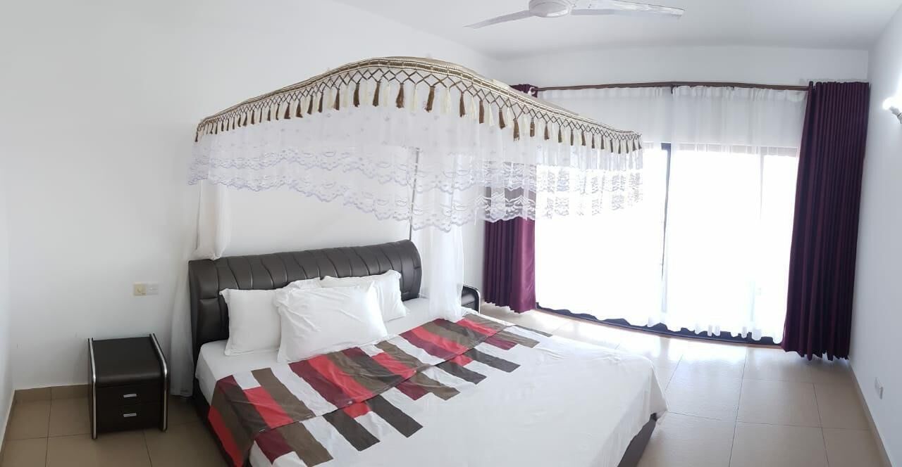 Un dormitorio con cama, mesita de noche, ventilador de techo y ventana con cortinas.