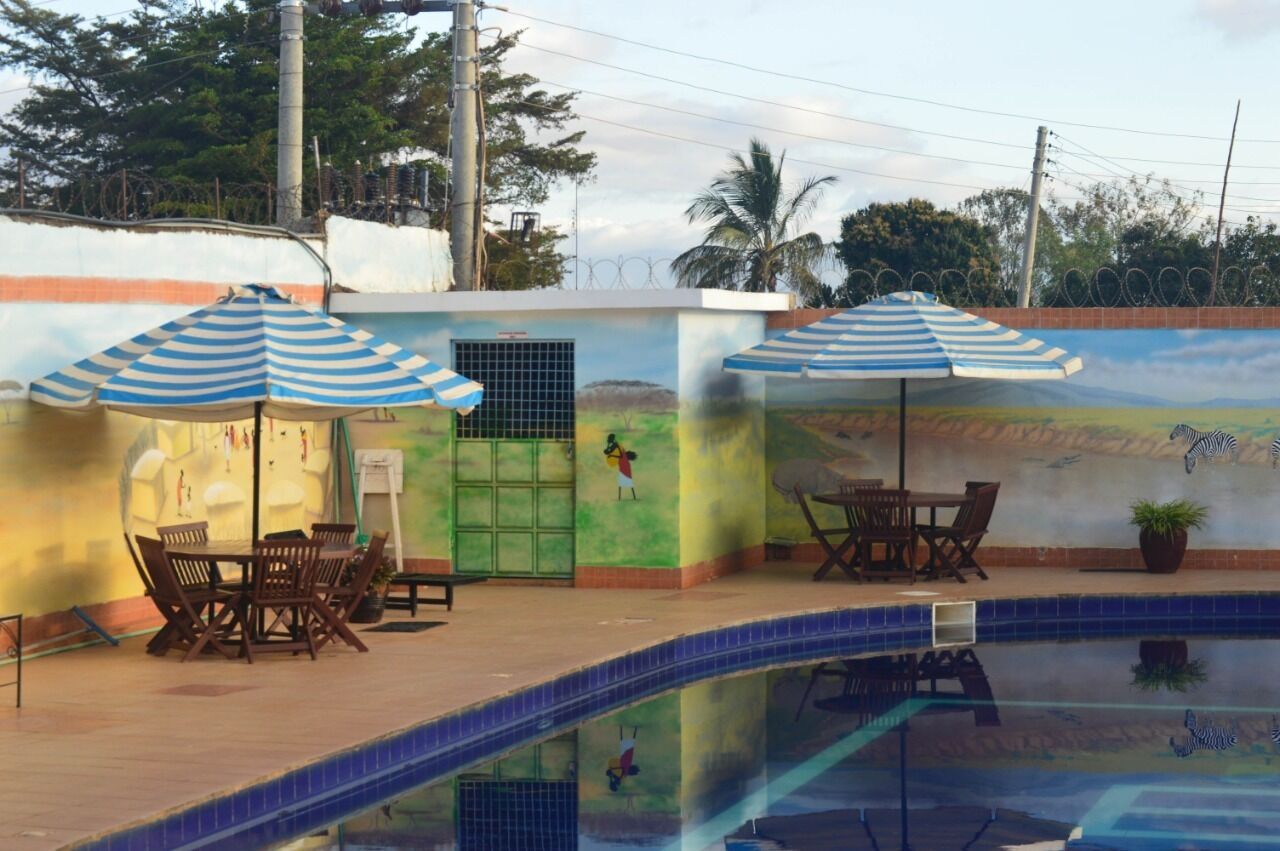 Zona de piscina con dos mesas y sillas bajo sombrillas a rayas, una puerta verde y un mural con cebras.