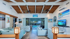 A spacious living area with two sofas, a coffee table, and a TV. There are ceiling fans, a large window with a view of the pool, and artwork on the walls.