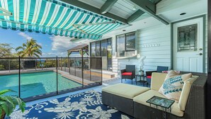 A covered outdoor area with a pool, a wicker sofa with cushions, and a dining table.