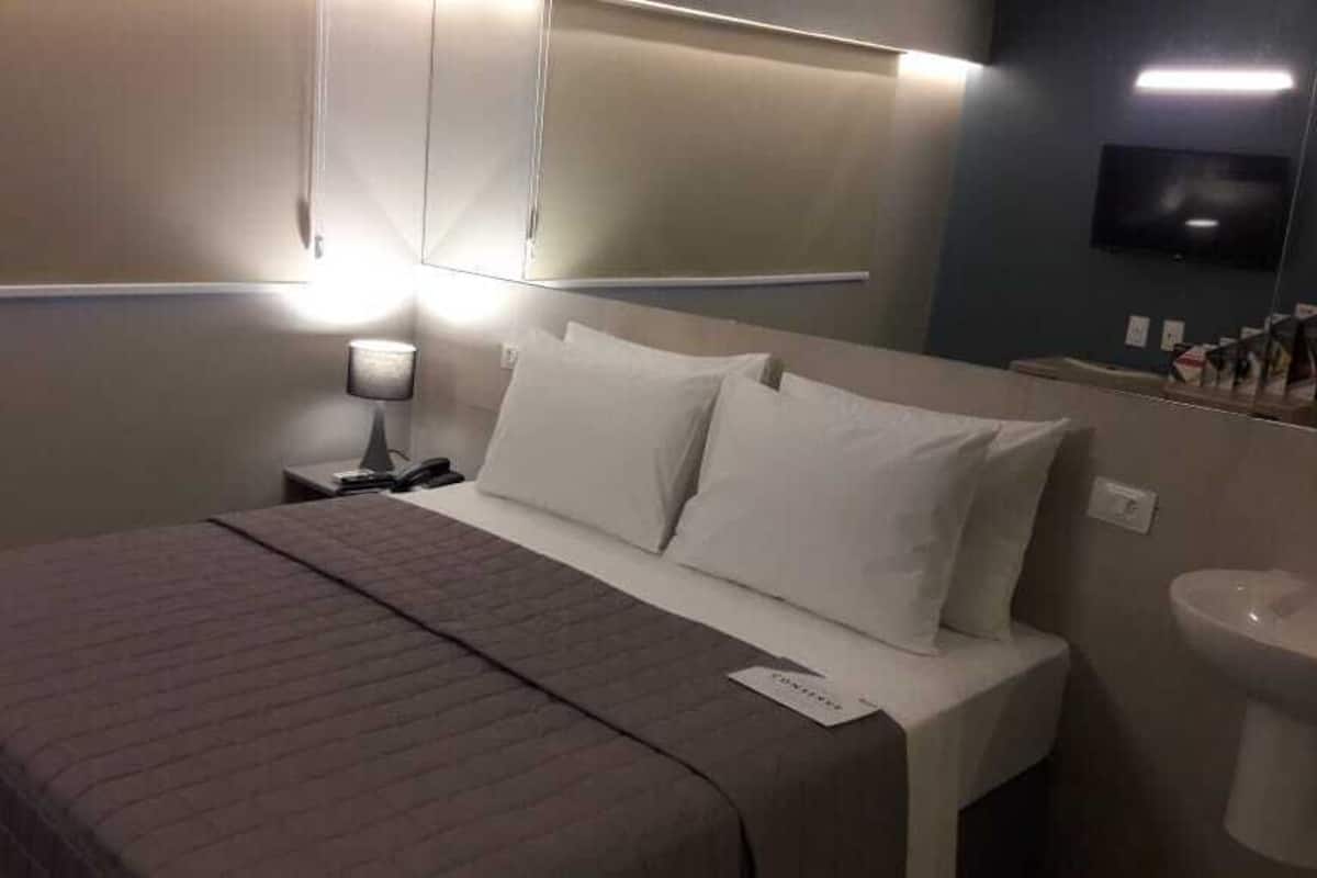 A hotel room with a bed, a nightstand with a lamp, a television mounted on the wall, and a toilet.
