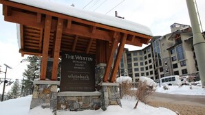 Un panneau enneigé indiquant l’emplacement du Westin Monache Resort Mammoth, un complexe hôtelier doté d’une base en pierre et d’une structure en bois.