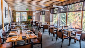 Un restaurant avec des poutres en bois apparentes au plafond, de grandes fenêtres offrant une vue sur le feuillage automnal et des tables soigneusement dressées.