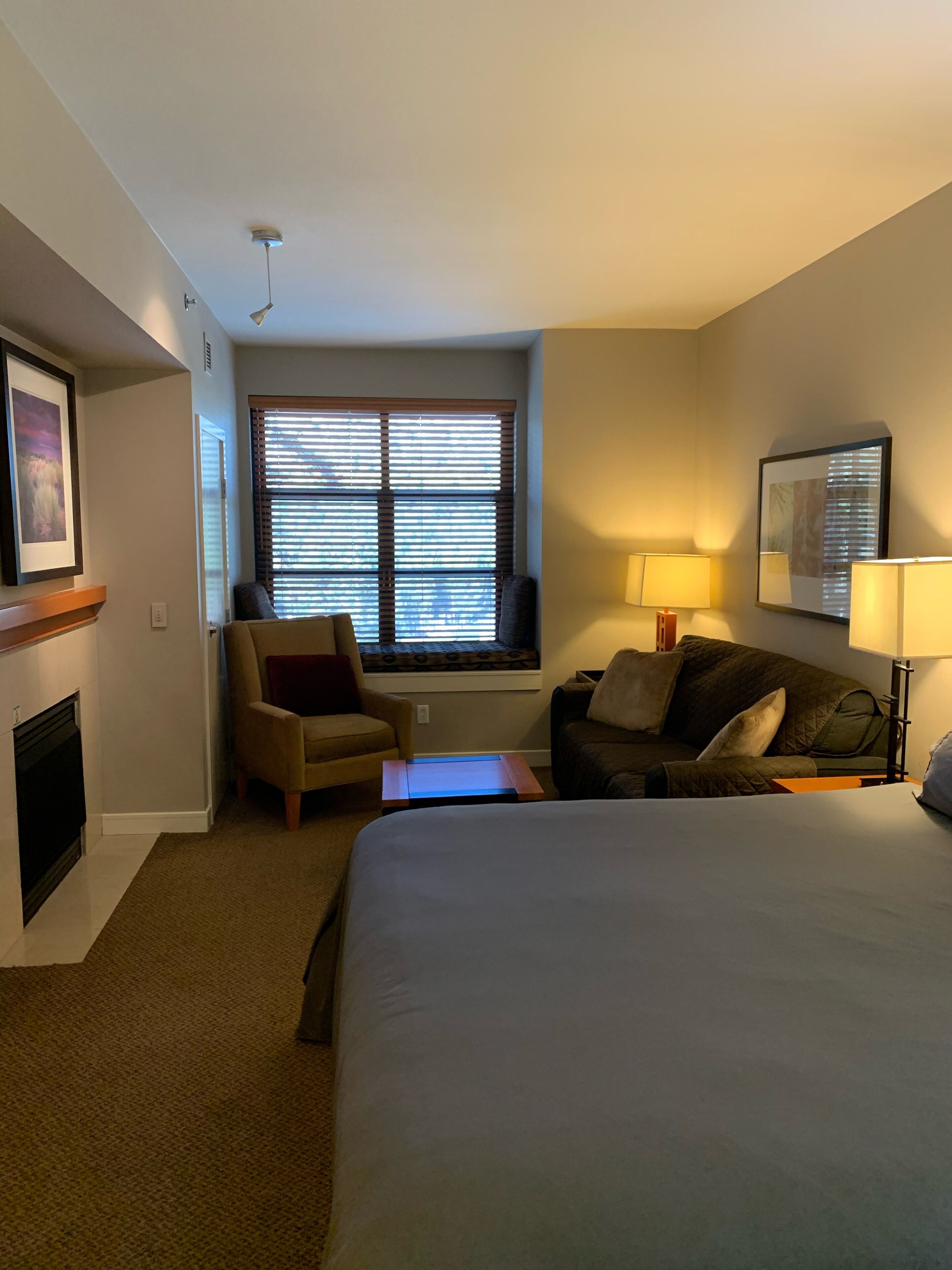 Habitación de hotel con cama, sofá, sillón, ventana con persianas, chimenea y lámpara de pared.