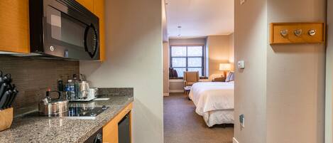 A compact hotel room with a kitchenette, a bed, a bedside table, and a window.